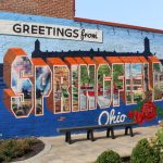 Colorful mural saying 'Greetings from Springfield, Ohio' with images of local land — ICE ankle monitors Haitian immigrants