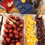 A school cafeteria table filled with fresh fruits like apples, oranges, and grapes, alongsid — SNAP cuts free school lunch