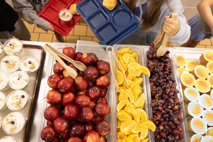 A school cafeteria table filled with fresh fruits like apples, oranges, and grapes, alongsid — SNAP cuts free school lunch