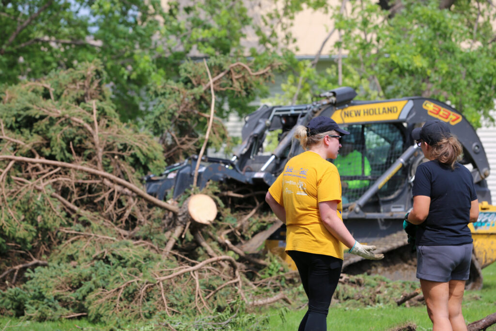 Two women discuss cleanup efforts near a tree debris pile and a construction vehicle after a storm, hi — FEMA disaster aid