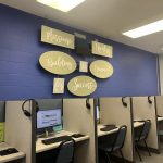 A room with cubicles containing computers and headsets, featuring a wall sign — women on parole mental health and housing