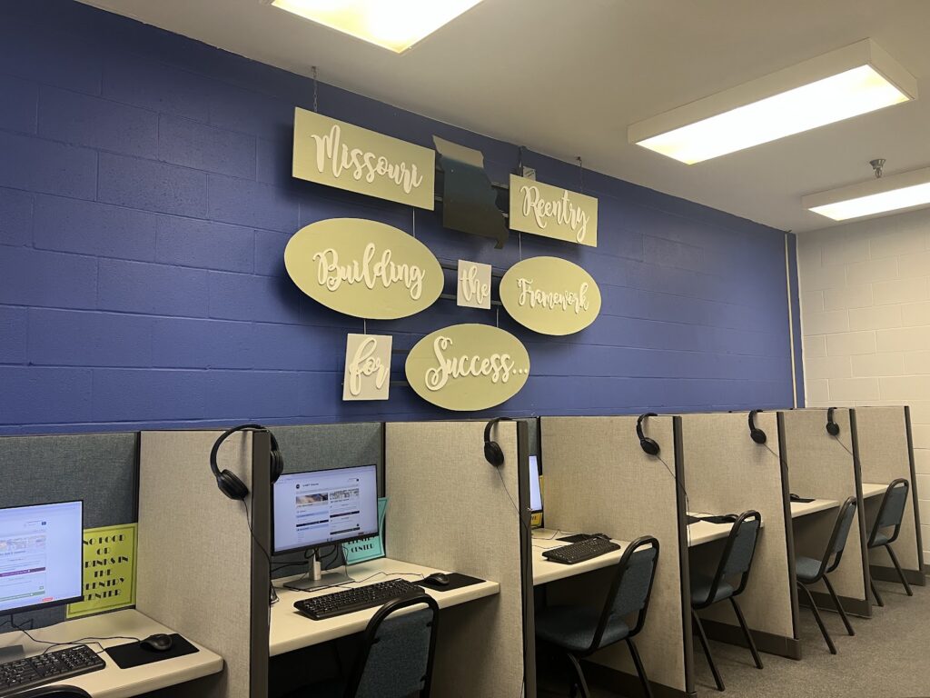 A room with cubicles containing computers and headsets, featuring a wall sign — women on parole mental health and housing