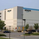 Amazon warehouse building with logo, surrounded by parking lot and trees, under a clear s — FTC Amazon advertising lawsuit