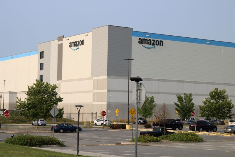 Amazon warehouse building with logo, surrounded by parking lot and trees, under a clear s — FTC Amazon advertising lawsuit