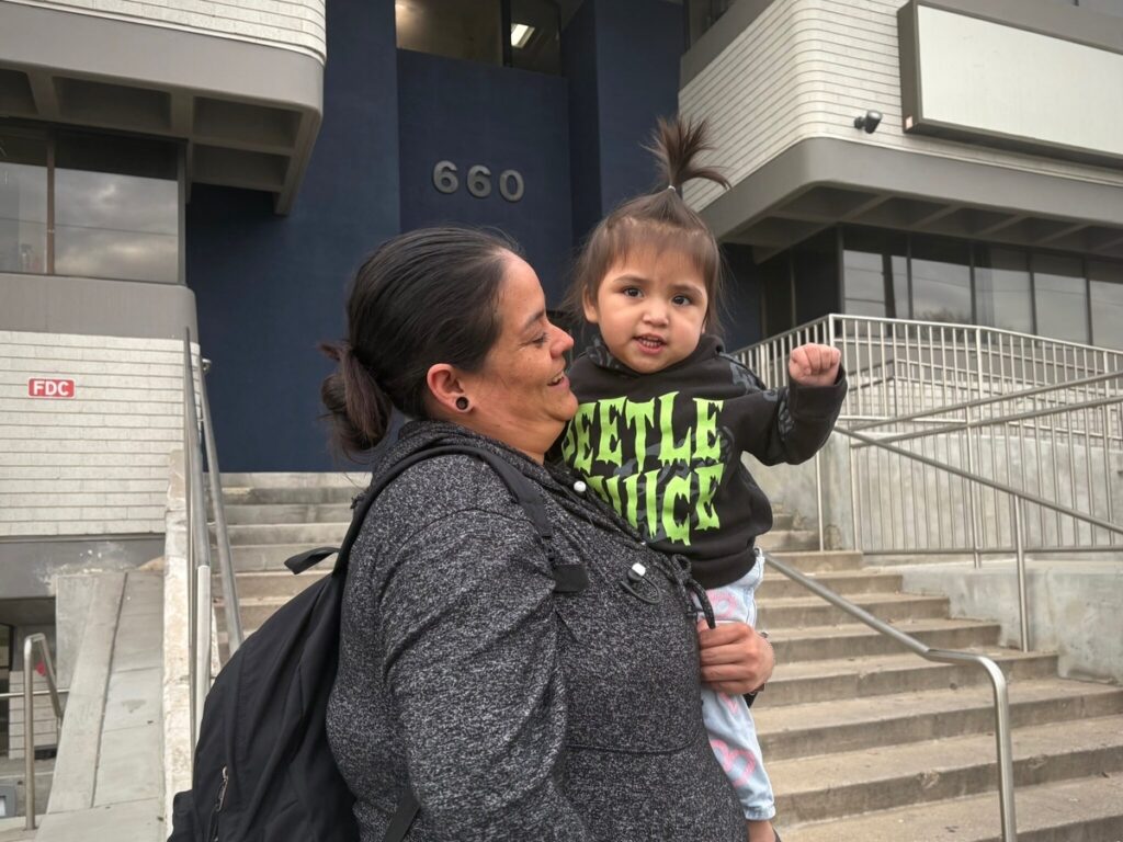 A woman holding a young child outside a building, both smiling, with the number 66 — trump green card restrictions lawsuit