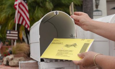 A hand placing a yellow mail-in ballot into a white mailbox, with an American flag in — USPS mail-in voting whistleblower