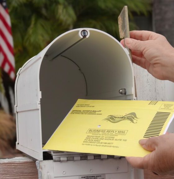 A hand placing a yellow mail-in ballot into a white mailbox, with an American flag in — USPS mail-in voting whistleblower