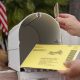 A hand placing a yellow mail-in ballot into a white mailbox, with an American flag in — USPS mail-in voting whistleblower