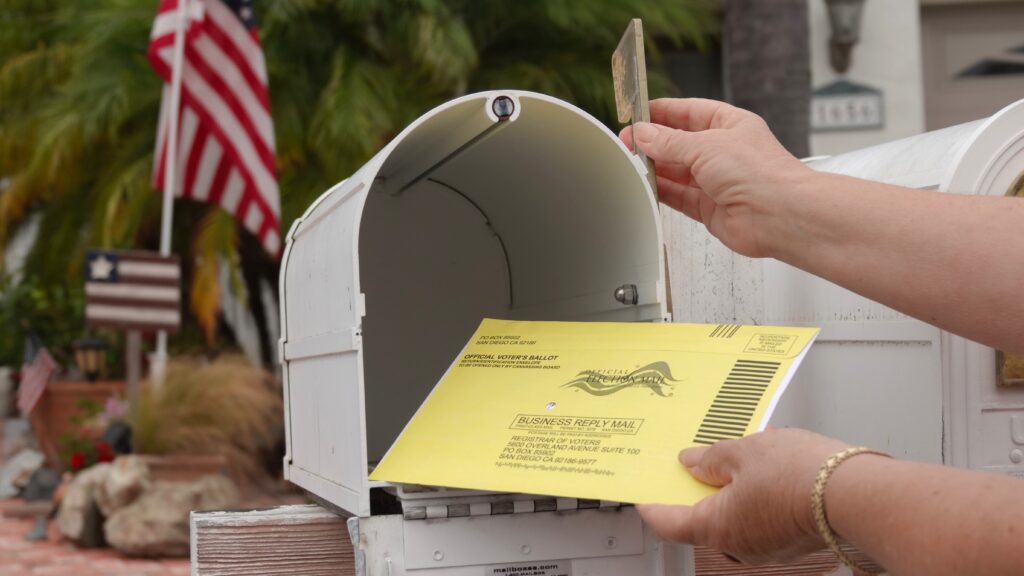 A hand placing a yellow mail-in ballot into a white mailbox, with an American flag in — USPS mail-in voting whistleblower