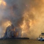 Smoke and flames rise from a wildfire near a body of water, with a boat in the foreground, il — wildfires impact on tribes