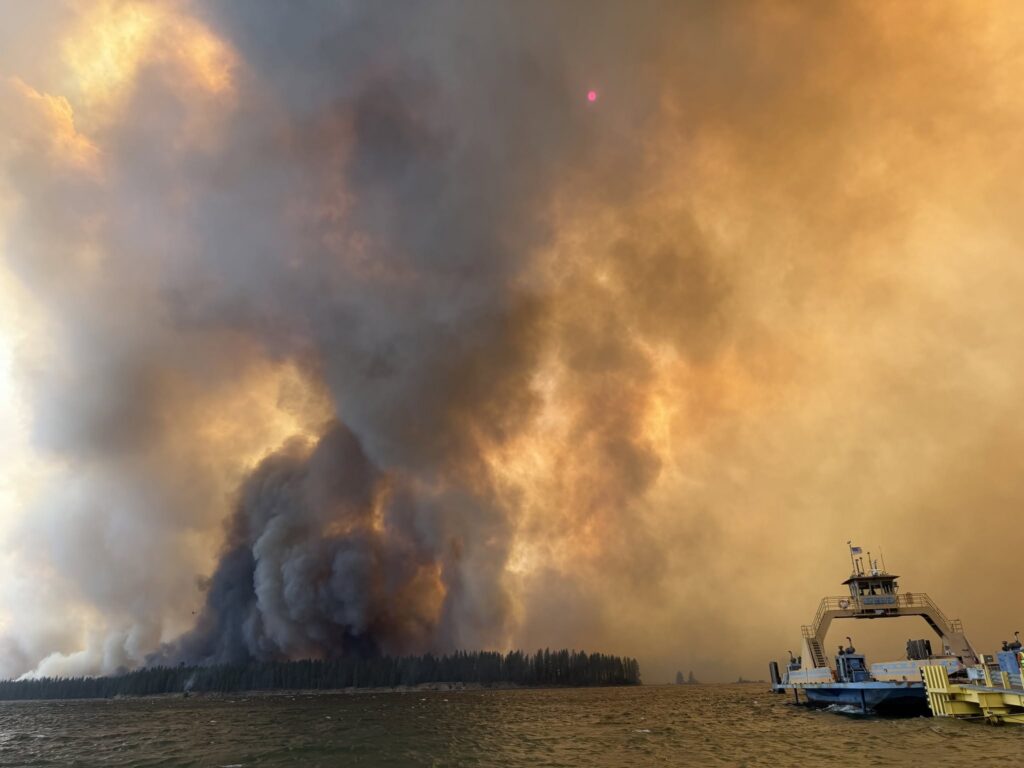 Smoke and flames rise from a wildfire near a body of water, with a boat in the foreground, il — wildfires impact on tribes