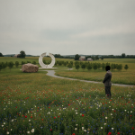 A person stands in a meadow of wildflowers near a memorial boulder and a circular monument. — Flight 93 25th anniversary