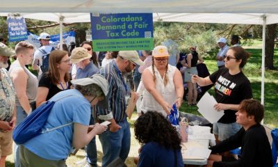 A diverse group of people gather under a tent, discussing tax initiatives and signing petition — state tax ballot measures