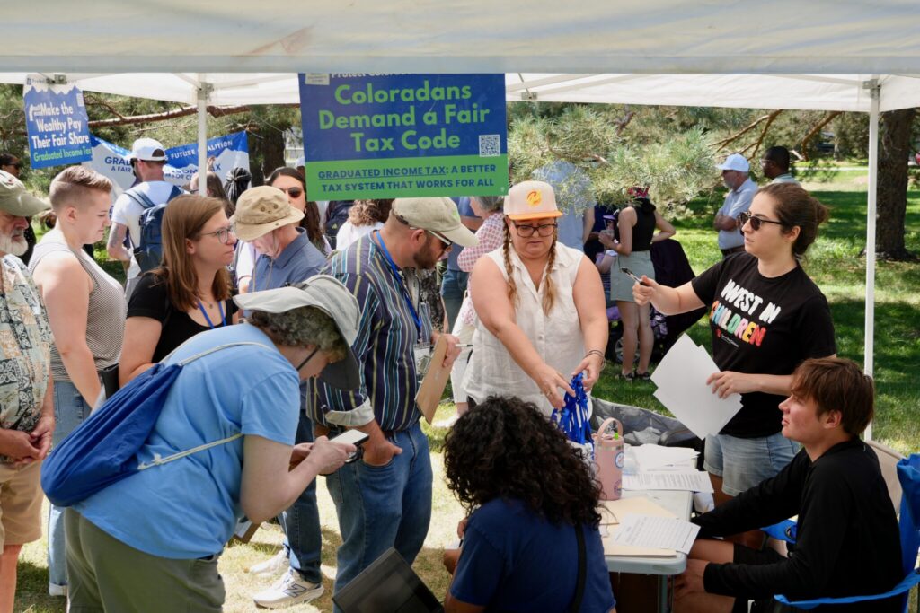 A diverse group of people gather under a tent, discussing tax initiatives and signing petition — state tax ballot measures