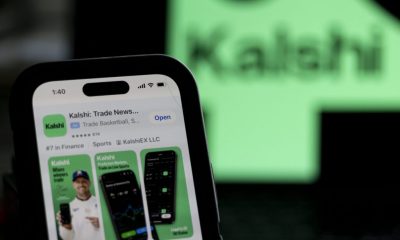 Utah sues Kalshi as a phone displays the Kalshi app listing in front of a blurred green Kalshi sign.