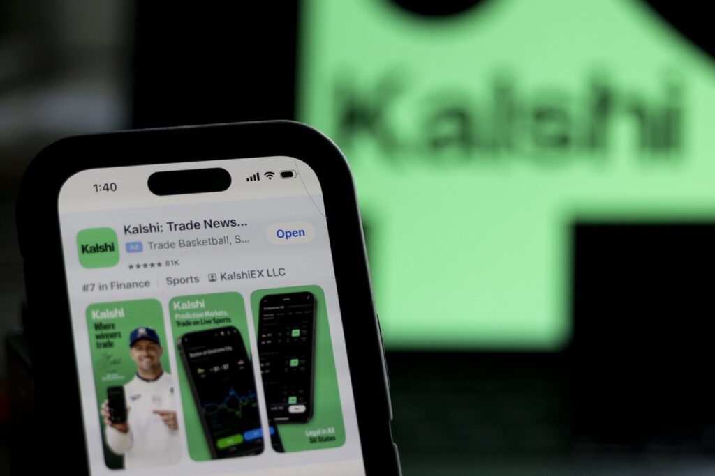 Utah sues Kalshi as a phone displays the Kalshi app listing in front of a blurred green Kalshi sign.