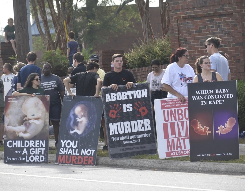 Criminal charges during pregnancy debate reflected in a sidewalk protest, with people holding anti-abortion signs and feta…