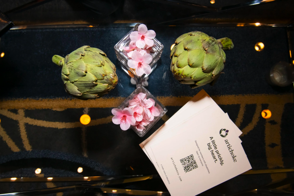 Artichokes, pink blossoms, and an Artichoke Credit Union business card arranged on a reflective table