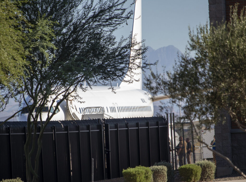 Trump deportation analysis: a white passenger plane behind a tall black fence, with people nearby