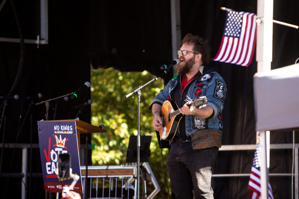 Musician performing at a No Kings movement rally with a guitar and an American flag in th — No Kings movement early voting