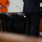A public defender stands at a courtroom table while a client in an orange jumpsuit waits in the — public defender shortage