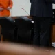 A public defender stands at a courtroom table while a client in an orange jumpsuit waits in the — public defender shortage