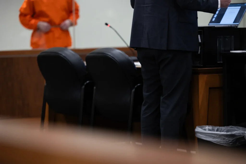 A public defender stands at a courtroom table while a client in an orange jumpsuit waits in the — public defender shortage