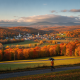 Bennington weather forecast over a sunlit autumn valley, with colorful hills, a village, and a person walking beneath a da…