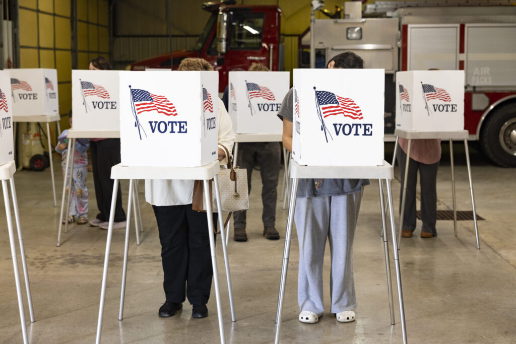 Trump DOJ election subpoenas story illustration: voters cast ballots at private voting booths marked with American flags.