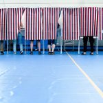 Voters standing behind striped privacy curtains in a polling place during an election. — Trump voter roll data lawsuit