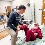 A caregiver helps an older man in a red outfit sit up in bed, illustrating the cost of caring for aging parents.