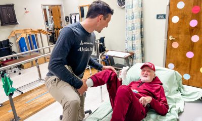 A caregiver helps an older man in a red outfit sit up in bed, illustrating the cost of caring for aging parents.