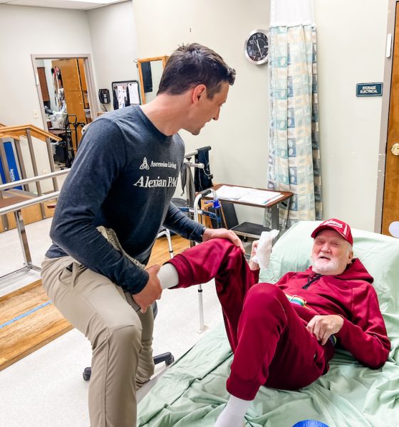 A caregiver helps an older man in a red outfit sit up in bed, illustrating the cost of caring for aging parents.