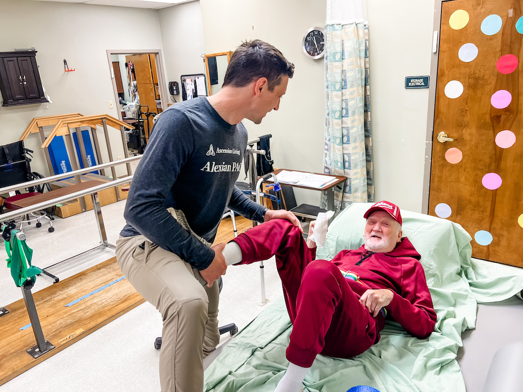 A caregiver helps an older man in a red outfit sit up in bed, illustrating the cost of caring for aging parents.
