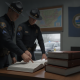 Two uniformed troopers review an open logbook beside stacked records at the Vermont State Police Shaftsbury Barracks.